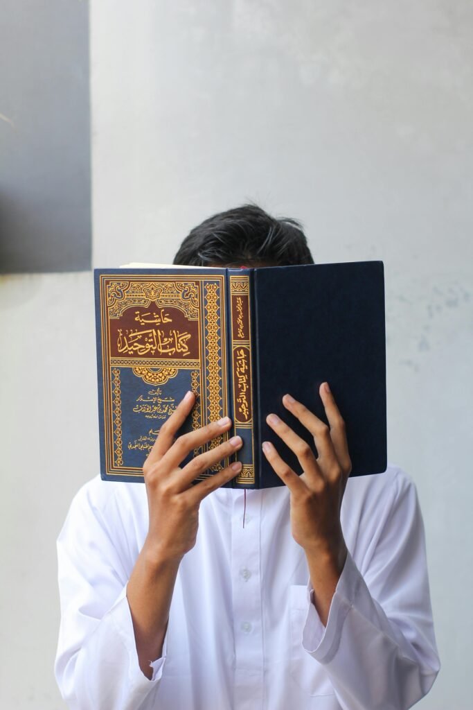 A person in white holding an Islamic book covering their face, focusing on religious study.